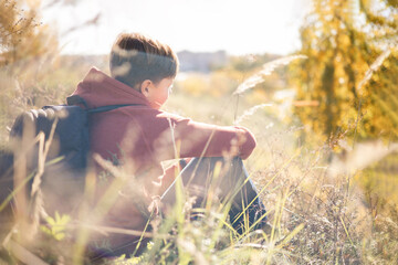 Autumn portrait of teenager boy sitting on grass. Young man with backpack resting relaxing,...