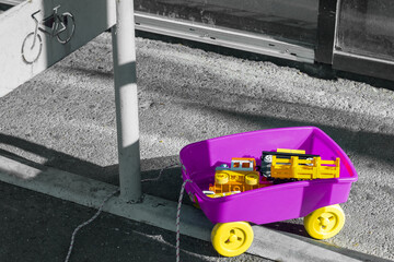 a bright purple children's cart with yellow wheels, abandoned forgotten toys in it; it stands despondently and alone near the store on a gray background; the theme of children's fears and growing up