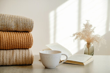 Stack of pastel color sweaters perfectly folded on the table. Open book, big cup of tea and a small vase. Fall winter season clothing, visible knitting pattern. Close up, copy space, background