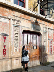 Fototapeta na wymiar Young candid millennial woman stands by shabby wall. Young brunette girl in a dress, leather jacket and sunglasses stands by old shabby chic wall and door. Old house building facade. Old music hall