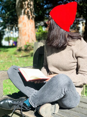 Fototapeta na wymiar Young candid woman in casual autumnal clothes siting on a wooden bench in park and reading red book. Early autumn. Wearing red hat and beige woollen sweater. Happy brunette, confident, generation z