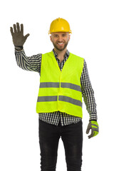 Smiling construction worker in reflective clothes is standing with hand raised. Stop gesture.
