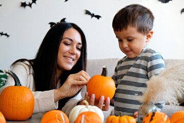 Young beautiful mother with her three year old son preparing decorations for halloween at home. Woman spending quality time with her child, carving pumpkins. Close up, copy space, background.