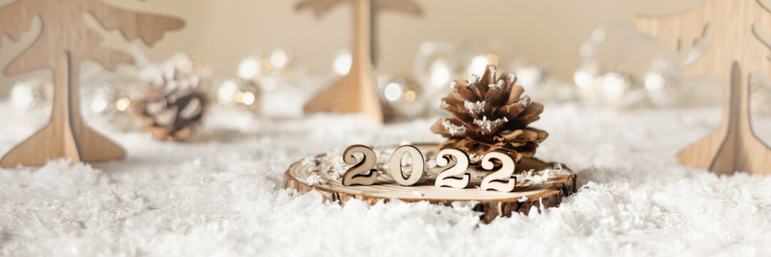 New year 2022. Numbers 2022 on wooden stand on beige pastel blurred background with decorative fir trees, snow and lights. Christmas greeting card. Banner