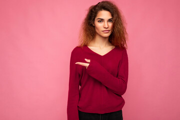 Photo of young beautiful brunette wavy woman with sincere emotions wearing casual pink sweater isolated over pink background with copy space and pointing at free space for mockup