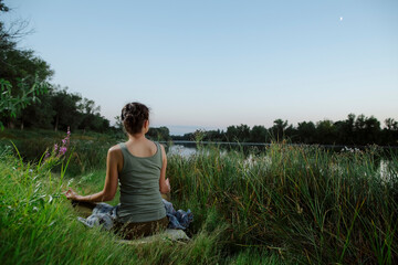 Yogin woman sits in a meditative position at sunsat and moon rise on the picturesque shore of lake