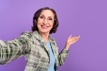 Photo of old attractive positive lady make selfie glasses invite welcome hand isolated on violet...