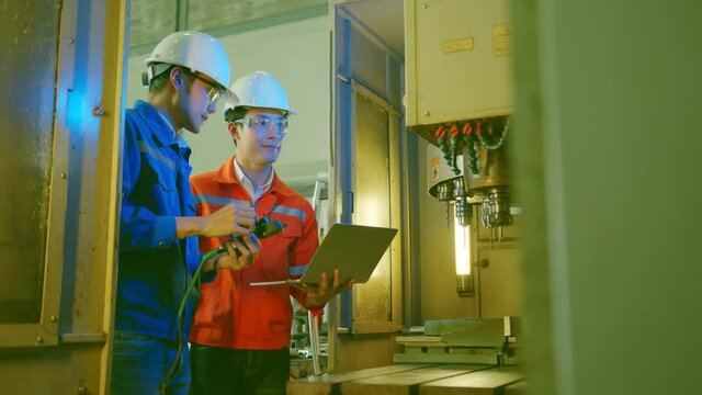 Co-working technician asian male engineer standing hand use laptop application result report discuss for quality control production line of cnc machine operate metal cutting work smart technology 