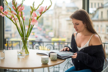 Cute caucasian girl reading a book or magazine at the cafe. Freelance, business concept. Morning routine.