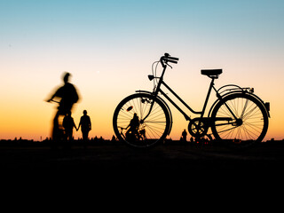 Fototapeta premium A silhouette of an old female bicycle against evening sky background standing on the field, people are walking around