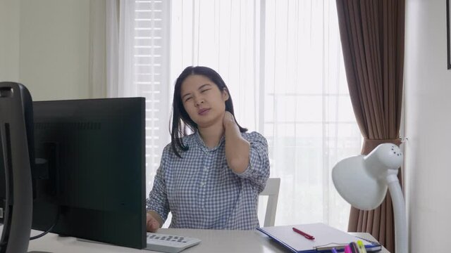 Medium shot portrait of a middle-aged, long hair Asian woman working or studying at home, using desktop computer, typing, having neck, shoulder or back pain. Office syndrome and muscles Inflammation.
