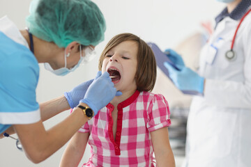 Otorhinolaryngologist doctor in protective medical mask examining throat of little girl using...