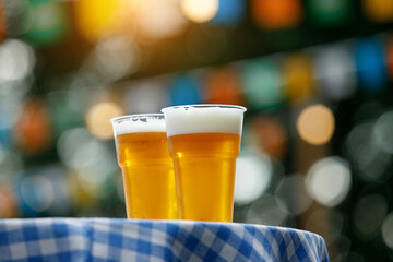 Two plastic glasses of beer on table with checkered tablecloth.
