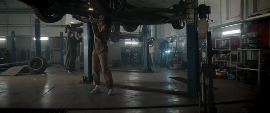Caucasian female mechanic repairing a car in a workshop, working under car bottom. Shot with 2x anamorphic lens