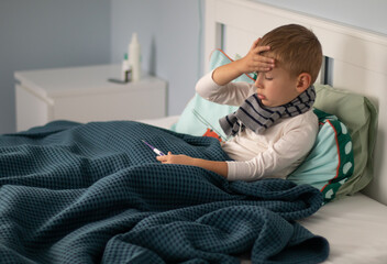 Obraz premium A little boy in a scarf is sick and lies in bed, holding a thermometer in his hands. Seasonal cold concept