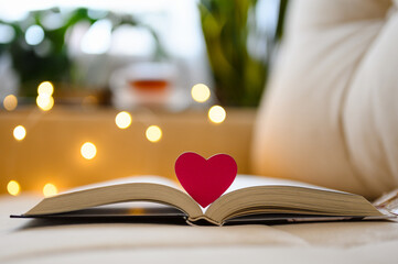 A small heart on an open book against the backdrop of a cozy home sofa and garland lights. Selective focus and narrow focus