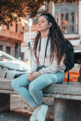 Adolescente joven realizando fotografías con su cámara de fotos en la ciudad descansando sentada en un banco junto a los taxis y el transporte publico para continuar con su ruta de viaje por España