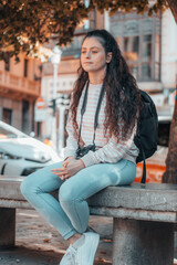 Chica joven fotógrafa reflexionando con su cámara de fotos en el pueblo descansando sentada en un banco junto a los taxis y el transporte publico para continuar con su ruta de viaje por España