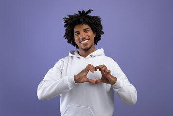 Kind black teenager shaping hands like heart near his chest, making love gesture on violet studio...
