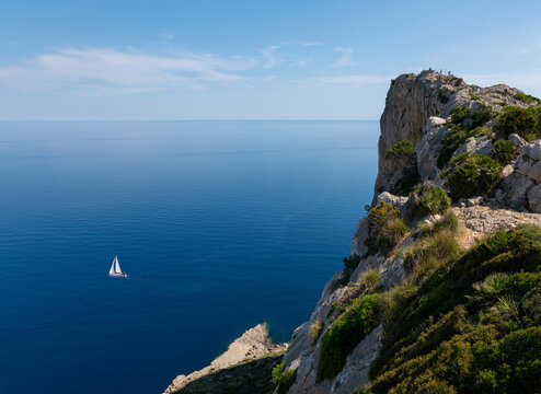 The steep cliffs of the Formentor peninsula in the east of the Mediterranean island of Mallorca. A sailing boat is sailing on the sea. The water is deep blue. Above is a viewing platform.