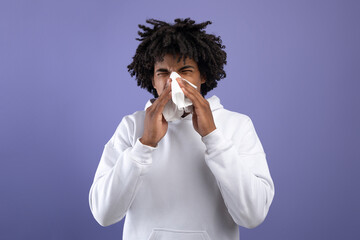 Cold and flu. Sick black teenager sneezing in paper tissue, suffering from rhinitis or sinusitis on violet background