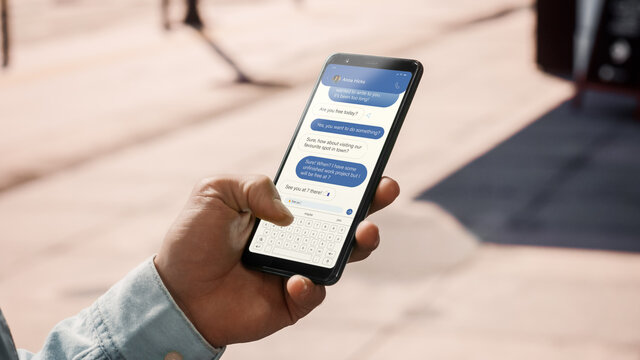 Close Up of a Young Man Using Smartphone For Sending Text Message to a Friend in Popular Social Media. Display is in Vertical Position. He is on a Street During a Sunny Day. He's Tapping the Screen.