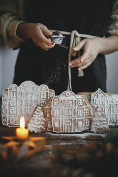 Woman in apron sprinkling sugar powder on christmas gingerbread houses on rustic wooden table. Atmospheric moody image. Christmas holiday preparation and traditions. Decorating cookies