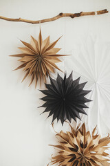 Stylish Christmas stars hanging on wooden branch on white wall. Modern festive scandinavian decor in room. Simple sweden craft, black and white paper stars.  Winter holiday preparation.