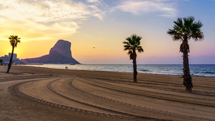 Sunset on the white sand beach with palm trees and big rock in the sea. Calpe Alicante.