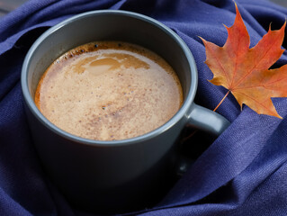 Latte coffee mug wrapped in blue fabric scarf, red maple leaf on table. Autumn hot drink, food concept, coffee shop flat lay close up