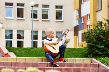 Little boy blond sits on steps in front of school and plays an acoustic guitar. Boy learns to play guitar and sing . It's stylish. Concept of studying music, guitar, hobby. Selective focus.