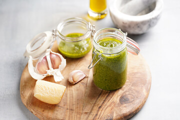 Several small glass mason jars with green italian basil sauce pesto on a round wooden tray with hard cheese pecorino and garlic cloves