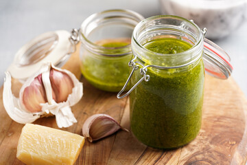 Several small glass mason jars with green italian basil sauce pesto on a round wooden tray with hard cheese pecorino and garlic cloves