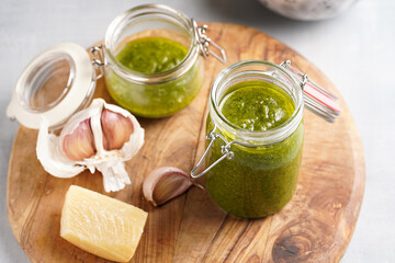 Several small glass mason jars with green italian basil sauce pesto on a round wooden tray with hard cheese pecorino and garlic cloves