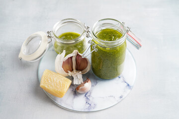 Several small glass mason jars with green italian basil sauce pesto on a round marble tray with hard cheese pecorino and garlic cloves