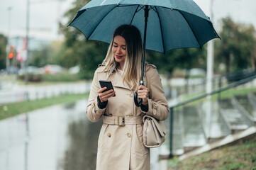 Businesswoman with umbrella using smartphone and walking down city street during rain