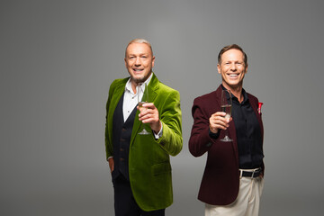 Smiling mature men in formal wear holding champagne isolated on grey