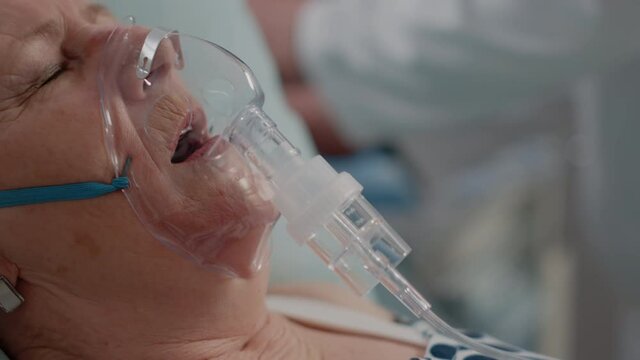 Close up of ill patient having respiratory problem in bed, while wearing oxygen mask against heavy breathing. Elder woman using medical equipment for healthcare. Hyperventilating adult