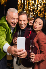 Blurred man with champagne taking selfie on smartphone with interracial friends during party on black background