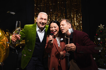 Happy mature interracial friends with champagne singing karaoke during new year party on black background