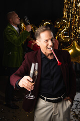 Smiling mature man with party horn and champagne looking at camera near blurred friend and festive decor on black background