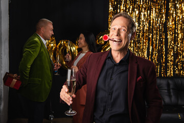 Cheerful mature man with party horn and champagne looking at camera near interracial friends and tinsel on black background