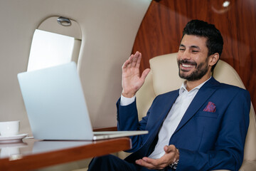 Handsome smiling Arabian businessman using laptop computer having video call flying luxury private jet.  Online meeting, video conference concept