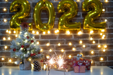 Merry Christmas and happy new year, defocused small Christmas tree on table with christmas presents and 2022 in golden inflatable numbers. Sparkling lights. Brick wall background