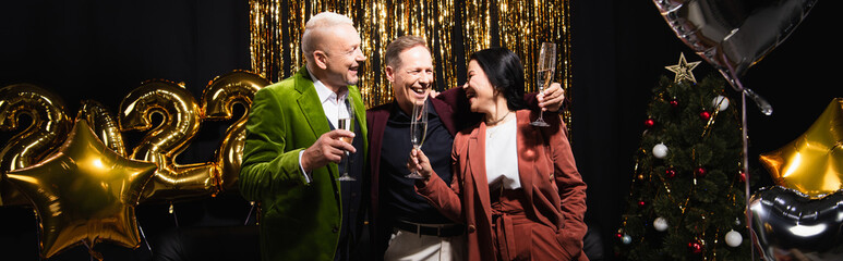 Positive interracial friends holding glasses of champagne near balloons, christmas tree on black background, banner