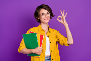 Photo of cute pretty young lady wear yellow shirt spectacles holding copybooks showing okey smiling isolated violet color background