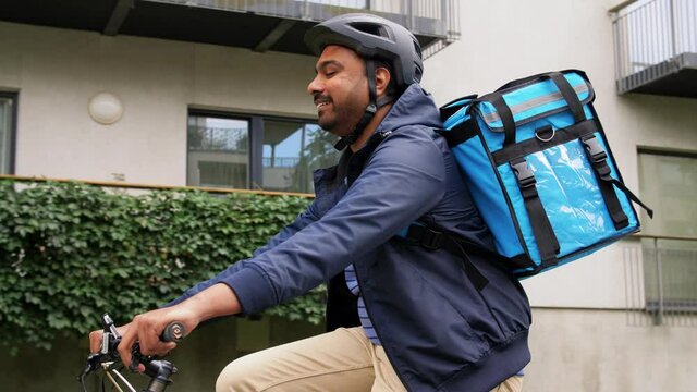 food shipping, profession and people concept - happy smiling indian delivery man in bike helmet with thermal insulated bag riding bicycle on city street