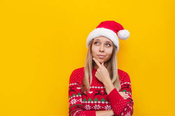 A young thoughtful blonde woman in a Christmas red deer warm sweater and a Santa's hat thinks about something looking away at empty copy space for text or design isolated on a color yellow background