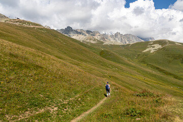 Obraz premium A woman hiking along a narrow pathway in high Caucasus mountains in Georgia. There are high glaciers in the back. Thick clouds above the sharp peaks. Lush pastures on the sides. Barren peaks.