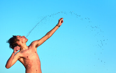 A closeup of the young naked man throwing water with the mouth standing on the background of blue sky.